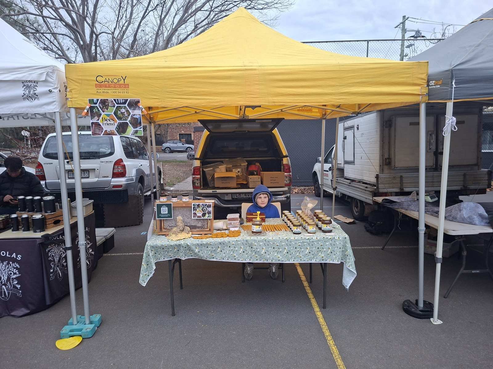 The bright yellow BeesFriend tent and honey stall at the Elwood Farmers Market, with jars of raw honey, honeycomb and beeswax candles on a bee-print tablecloth