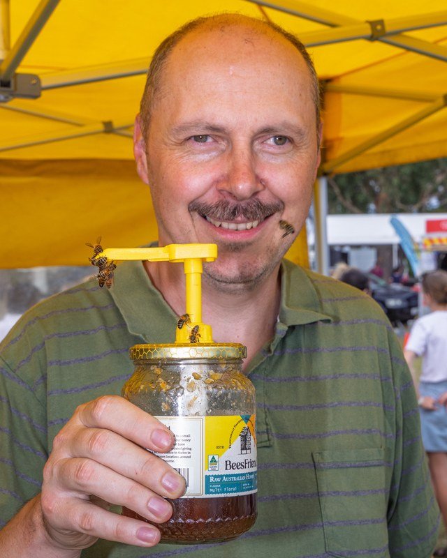 Laszlo Kun of BeesFriend holding a jar of raw honey at a market stall, with bees on the dispenser