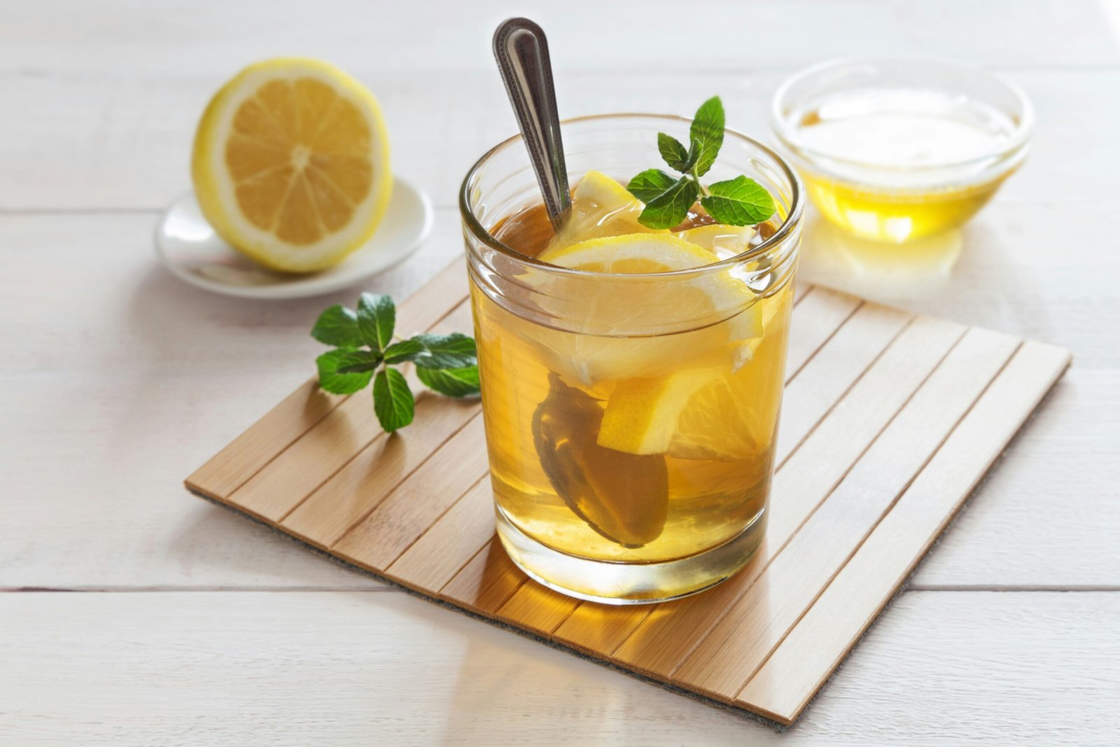 A glass of warm honey water with lemon slices and mint, a bowl of honey behind